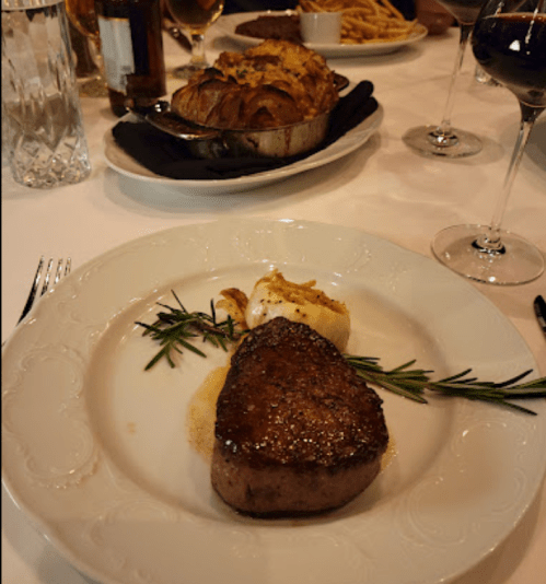 A plate with a seared steak garnished with rosemary, accompanied by creamy mashed potatoes and a side dish in the background.
