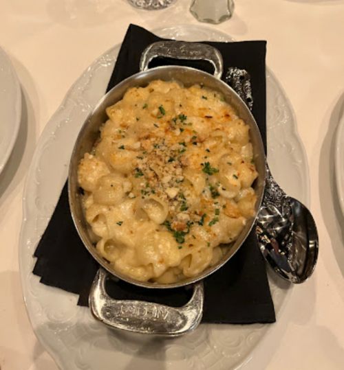 A serving of baked macaroni and cheese in a silver dish, topped with breadcrumbs and parsley, on a black napkin.