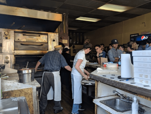 A busy kitchen scene with chefs preparing food, customers waiting, and pizza boxes stacked on a counter.