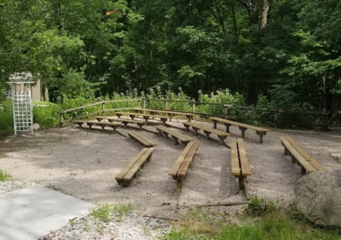 An outdoor amphitheater with wooden benches arranged in a semi-circle, surrounded by trees and greenery.