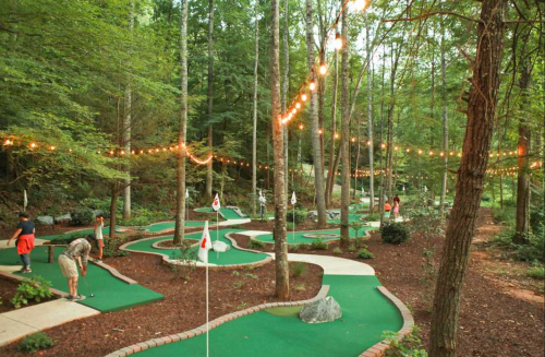 A mini-golf course winding through a wooded area, with players and string lights illuminating the scene.