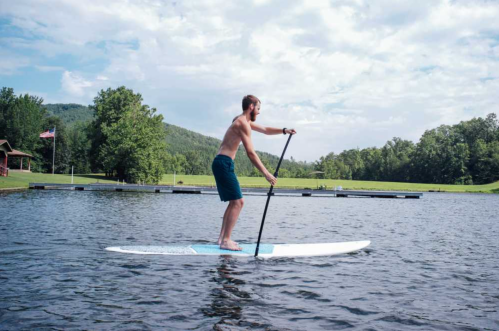 A man stands on a paddleboard in a calm lake, surrounded by trees and a cloudy sky.