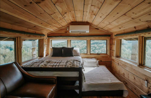 Cozy wooden interior of a cabin bedroom with a bed, a mattress on the floor, and large windows showcasing a scenic view.