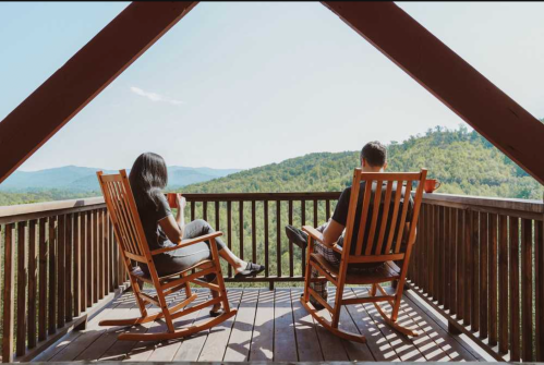 Two people sit in rocking chairs on a balcony, overlooking a scenic mountain view on a sunny day.