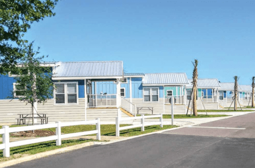 Row of modern blue and gray modular buildings with metal roofs, surrounded by greenery and a clear blue sky.