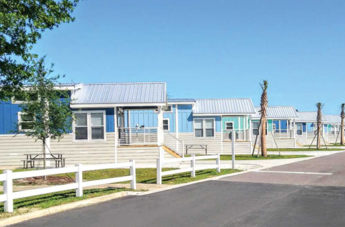 Row of modern blue and gray modular buildings with metal roofs, surrounded by greenery and a clear blue sky.