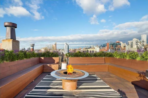 Rooftop terrace with a fire pit, surrounded by greenery and city skyline under a blue sky with clouds.