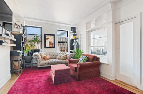 Cozy living room with a tufted sofa, armchair, colorful cushions, plants, and a vibrant red rug. Bright natural light.