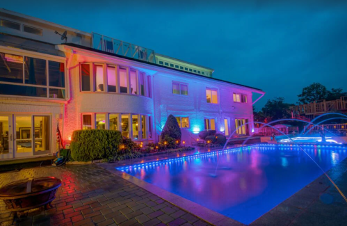 A modern home at dusk, featuring colorful lights and a pool with fountains, surrounded by landscaped greenery.