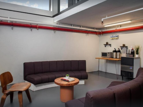 A modern lounge area with dark sofas, a wooden table, and kitchen appliances against a minimalist backdrop.