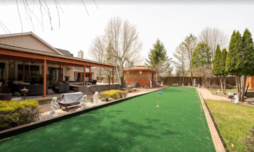 A backyard with a green lawn, patio area, and trees, featuring a bocce ball court and outdoor seating.