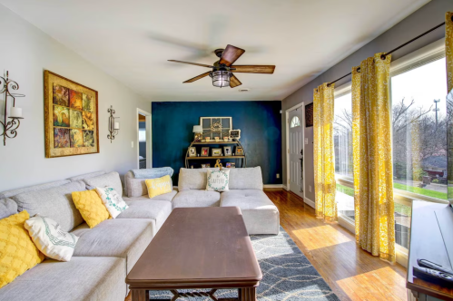Bright living room with a gray sectional sofa, yellow accents, wooden floor, and large windows letting in natural light.