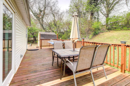 A wooden deck with a table and chairs, featuring an umbrella, surrounded by greenery and a shed in the background.