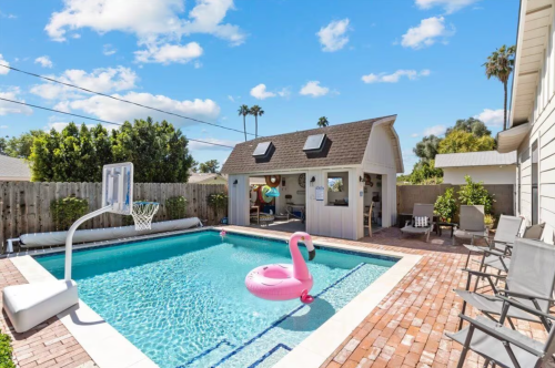 A sunny backyard with a pool, pink flamingo float, basketball hoop, and a cozy shed surrounded by greenery.