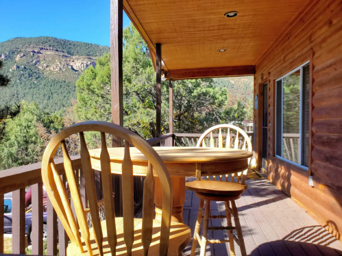 A wooden deck with a round table and chairs, surrounded by trees and mountains under a clear blue sky.