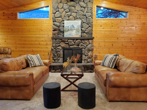 Cozy living room with stone fireplace, wooden walls, two leather sofas, and a coffee table with decorative items.