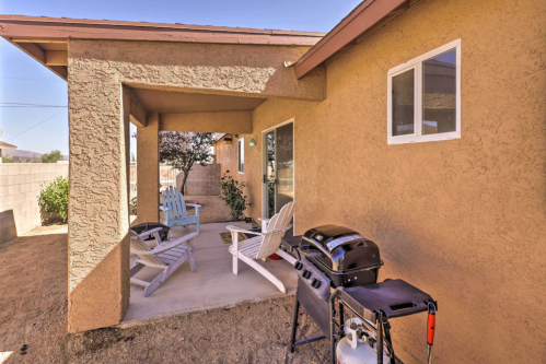 A patio area with two white chairs, a small table, and a grill beside a beige house in a sunny outdoor setting.