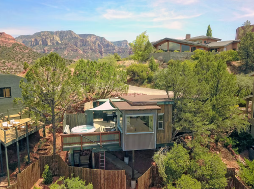 A modern cabin nestled among trees, featuring a hot tub and stunning mountain views in the background.