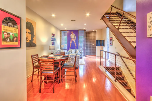 A modern living space with a dining area, staircase, and vibrant artwork on the walls. Warm wood floors enhance the ambiance.