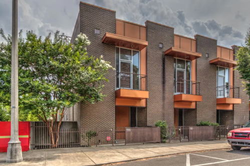 Modern brick townhouses with balconies, surrounded by greenery, under a cloudy sky.