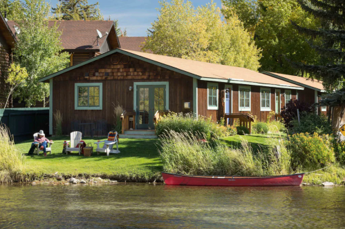 A cozy wooden house by a lake, with chairs on the lawn and a red canoe tied to the shore. Trees in the background.