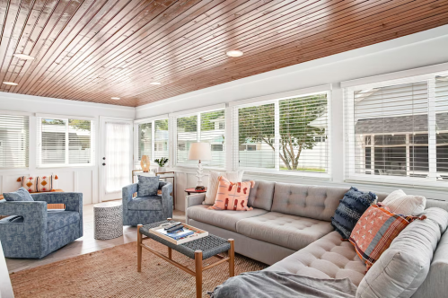 Bright sunroom with wooden ceiling, large windows, cozy seating, and decorative pillows, creating a welcoming atmosphere.
