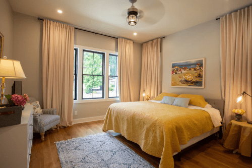 Cozy bedroom with a yellow bedspread, large windows, soft curtains, and a decorative painting on the wall.