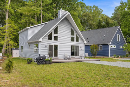 A modern two-story house with a sloped roof, surrounded by greenery and a gravel path, featuring a deck and outdoor seating.