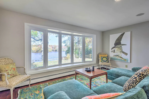 Cozy living room with a view of a lake, featuring a green sofa, a wooden table, and a large bird painting on the wall.