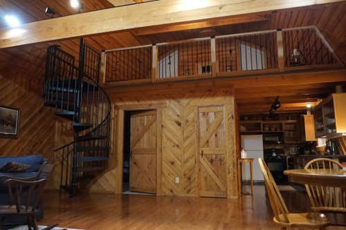 Cozy wooden interior of a cabin with a spiral staircase, open kitchen, and a loft area above. Warm lighting enhances the ambiance.