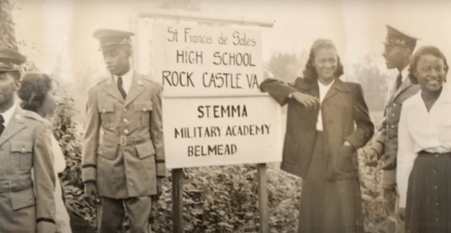 Historical photo of a group of people in military uniforms and civilian clothes by a sign for St. Francis de Sales High School.