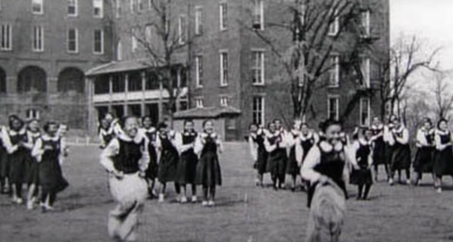 Black and white photo of a group of girls in school uniforms playing outside in front of a large brick building.