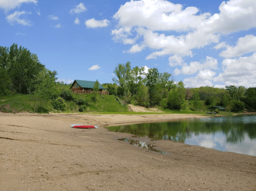 A sandy beach by a calm lake, with green trees and a cabin in the background under a partly cloudy sky.
