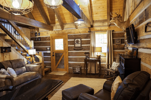 Cozy cabin interior with wooden beams, a staircase, leather sofas, and rustic decor, including a mounted deer head.