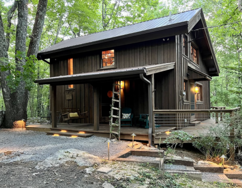 A rustic wooden cabin surrounded by trees, featuring a porch, ladder, and warm lights glowing from the windows.