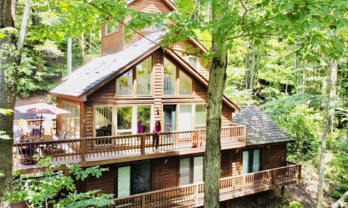 A large wooden cabin surrounded by lush green trees, featuring multiple windows and a spacious deck.