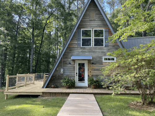 A charming A-frame cabin surrounded by trees, featuring a wooden deck and colorful plants at the entrance.