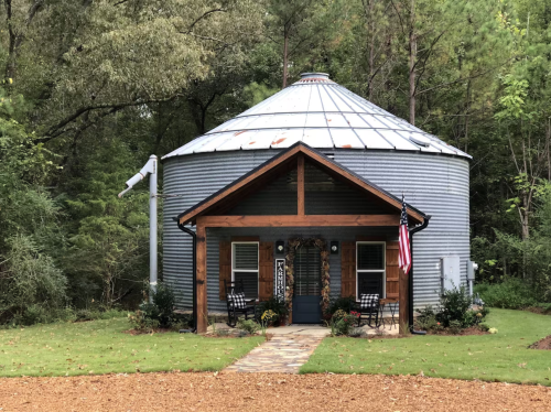 A round, metal-clad house with a wooden porch, surrounded by greenery and a gravel path. American flag displayed.