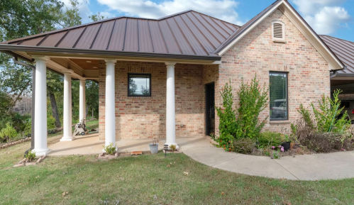 A brick house with a metal roof, featuring white columns and a landscaped yard under a blue sky.