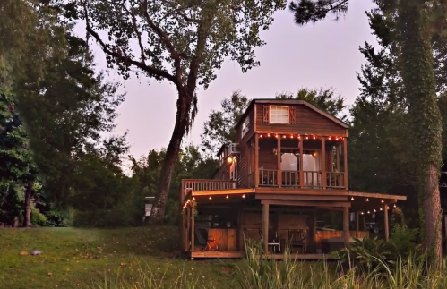 A charming two-story wooden house with string lights, surrounded by trees and greenery at dusk.