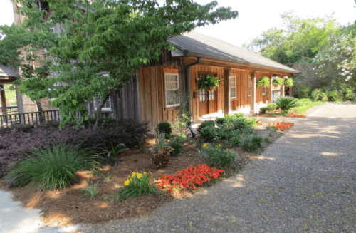 A charming wooden building surrounded by colorful flower beds and greenery on a sunny day.