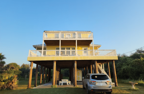 A yellow house on stilts with a spacious deck, surrounded by greenery and a parked car in the foreground.