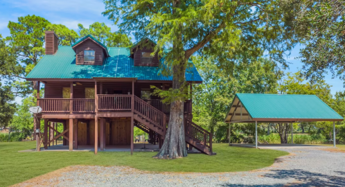A two-story wooden house with a green metal roof, surrounded by trees and a covered parking area.