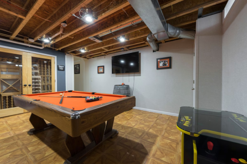 A basement game room featuring a pool table, TV, and arcade machine, with wooden beams and framed pictures on the walls.