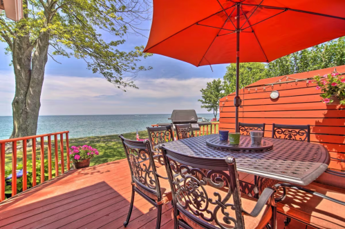 A scenic deck with a table and chairs, an orange umbrella, and a view of the lake and trees in the background.