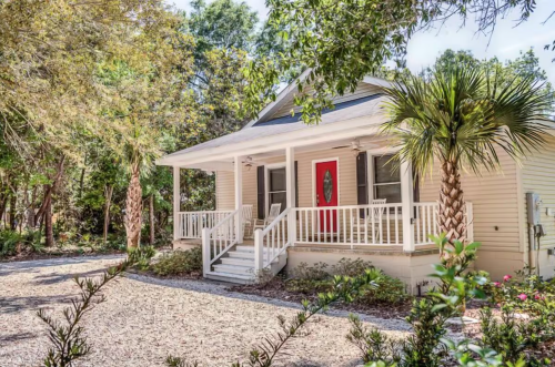 A charming yellow house with a red door, surrounded by greenery and palm trees, set on a gravel path.