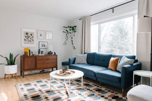 A cozy living room featuring a blue sofa, wooden furniture, plants, and a patterned rug under natural light.