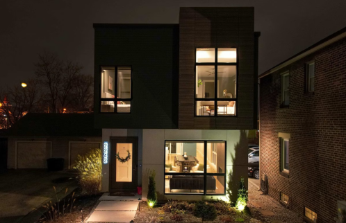 Modern two-story house at night, featuring large windows and warm interior lighting, surrounded by landscaping.