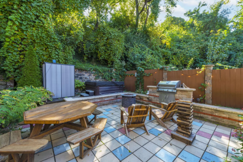 A cozy outdoor patio with a wooden table, chairs, a grill, and lush greenery in the background.