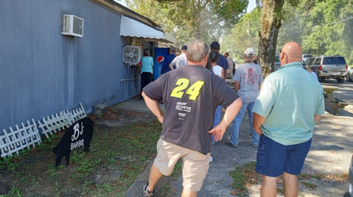 A group of people waiting in line outside a barbecue restaurant on a sunny day.